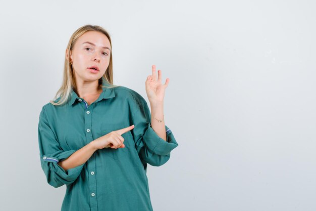 Chica rubia mostrando el signo de ok mientras lo señala con una blusa verde y se ve seria