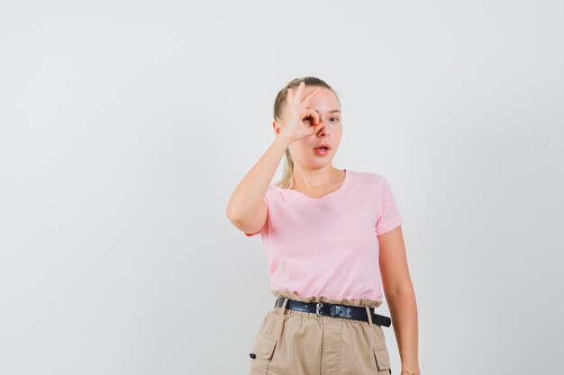 Chica rubia mostrando signo ok en el ojo en camiseta, pantalón, vista frontal.