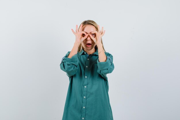 Chica rubia mostrando signos de ok en los ojos en blusa verde y mirando divertido