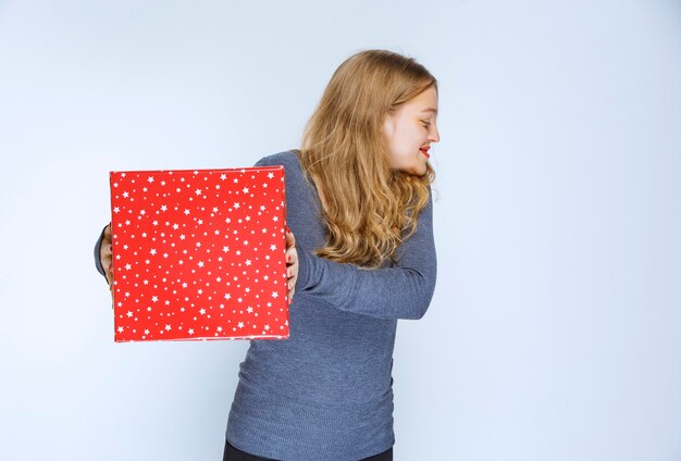 Chica rubia ofreciendo una caja de regalo roja a alguien.