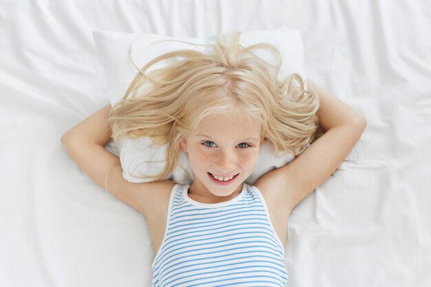 Chica rubia pecosa acostada en la cama, con expresión complacida mientras se regocija en el comienzo del nuevo día, teniendo fines de semana, sin ir a la escuela. Niño feliz sonriente que tiene buena relajación en cama cómoda