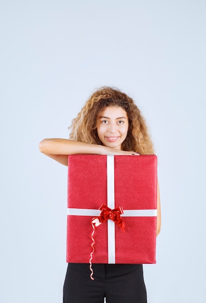 Chica rubia con pelos rizados sosteniendo una caja de regalo roja y sintiéndose positiva.