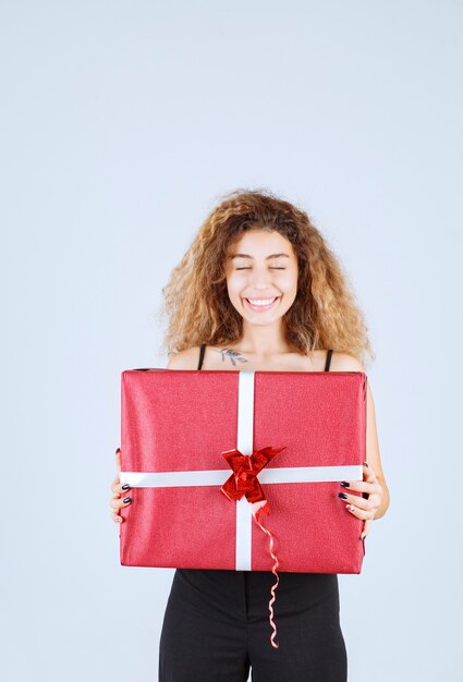 Chica rubia con pelos rizados sosteniendo una caja de regalo roja y sintiéndose positiva.