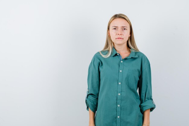 Chica rubia de pie con la espalda recta, mordiendo los labios y posando a la cámara en blusa verde y mirando seria.