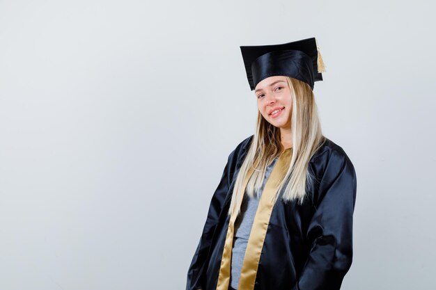 Chica rubia de pie con la espalda recta y posando a la cámara en toga y gorra de graduación y mirando feliz.