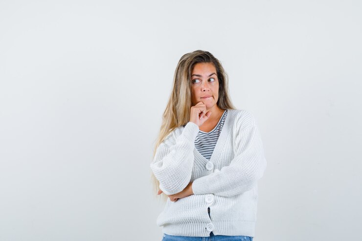 Foto gratuito chica rubia de pie en pose de pensamiento en camiseta a rayas, cárdigan blanco y pantalones de mezclilla y mirando pensativo. vista frontal.