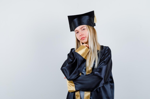 Chica rubia de pie en pose de pensamiento en toga y birrete de graduación y mirando pensativo