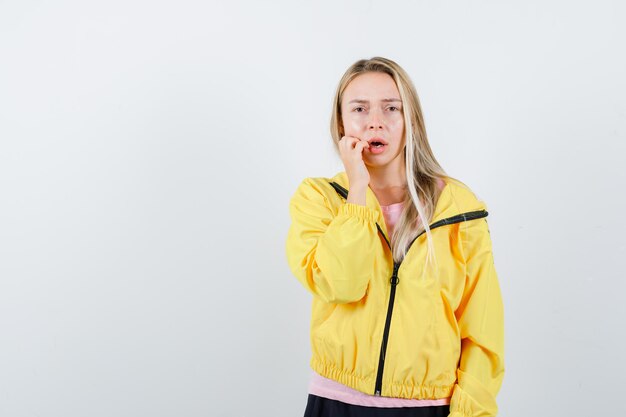 Chica rubia poniendo la mano cerca de la boca, manteniendo la boca abierta en camiseta rosa y chaqueta amarilla y mirando sorprendida