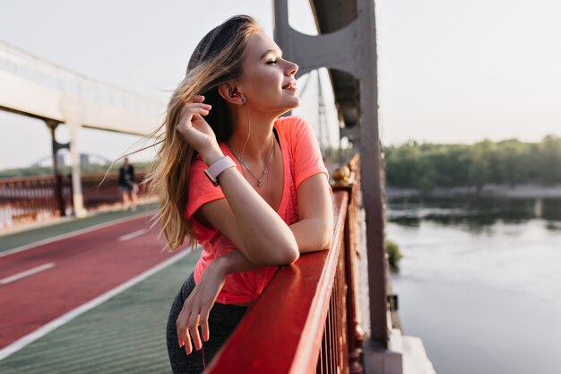 Chica rubia positiva escuchando música después del entrenamiento. Hermosa mujer caucásica relajante en una buena mañana de verano.