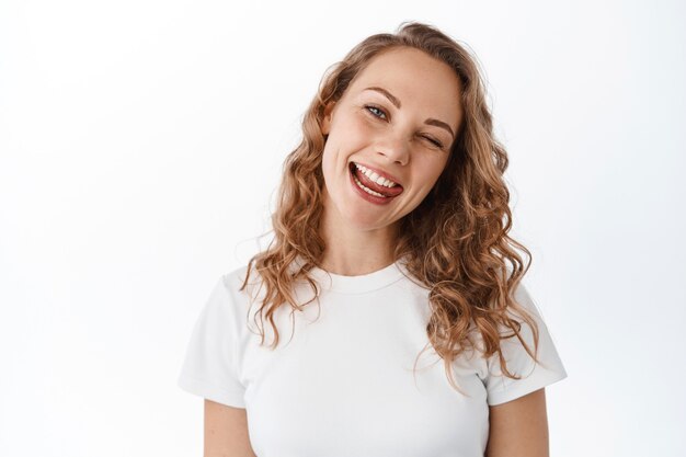 Chica rubia positiva guiñando un ojo, mostrando la lengua y sonriendo feliz, manteniéndose alegre y optimista, mirando con alegría al frente, de pie en camiseta contra la pared blanca