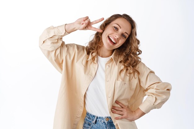 Chica rubia positiva mostrando el signo v de paz, sonriendo e inclinando la cabeza con expresión de cara feliz, de pie contra el fondo blanco.