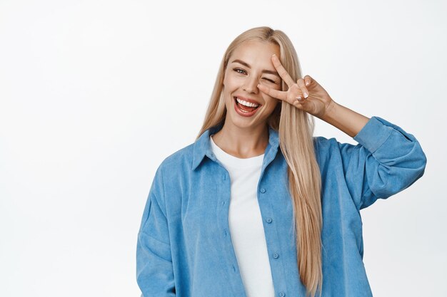 Una chica rubia positiva muestra un gesto de paz contra el ojo y guiña un ojo felizmente sonriendo con dientes blancos a la cámara de pie contra el fondo del estudio