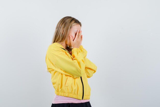 Chica rubia que cubre la cara con las manos en la chaqueta amarilla y parece deprimido