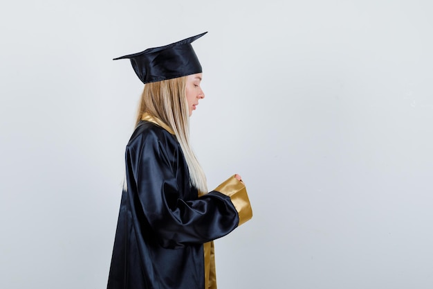 Chica rubia que finge sostener algo con ambas manos en toga y gorra de graduación y se ve linda.