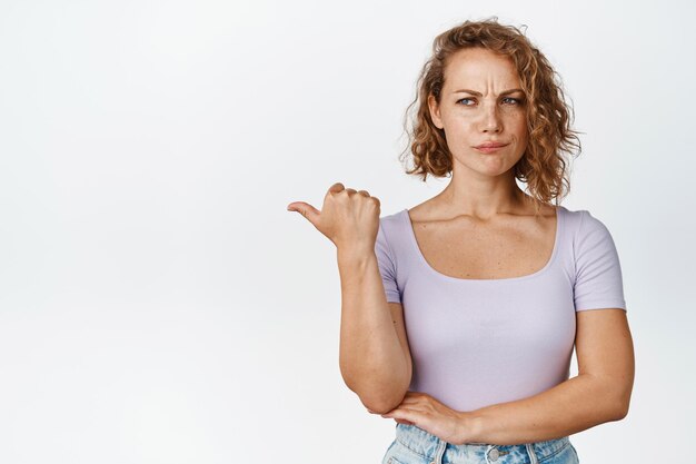 Chica rubia renuente frunciendo el ceño, señalando y mirando a la izquierda con expresión decepcionada, parada en camiseta sobre fondo blanco.