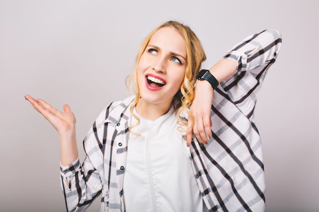 Chica rubia rizada en ropa elegante con mirada de disgusto en la cara escucha el reloj de pulsera roto. Close-up retrato de mujer joven decepcionada con lindo peinado con camisa a rayas, agitando las manos