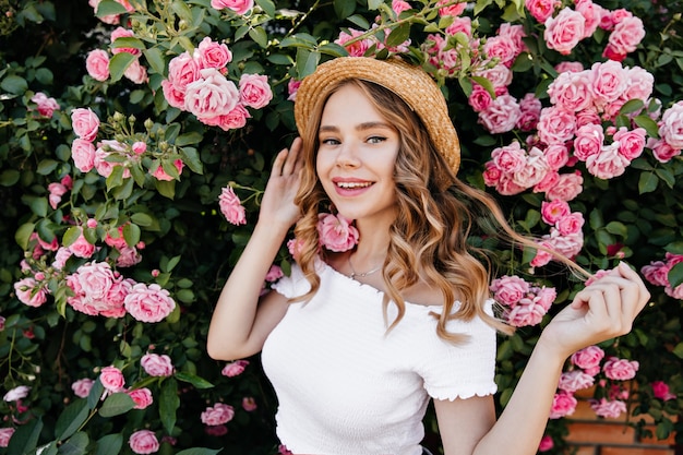 Chica rubia romántica posando con una sonrisa delante de hermosas flores. Retrato al aire libre de mujer rizada alegre jugando con su cabello en el jardín.