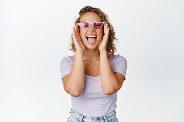 Chica rubia salvaje y libre divirtiéndose, usando gafas de sol, mostrando la lengua y gritando despreocupada, de pie feliz contra el fondo blanco