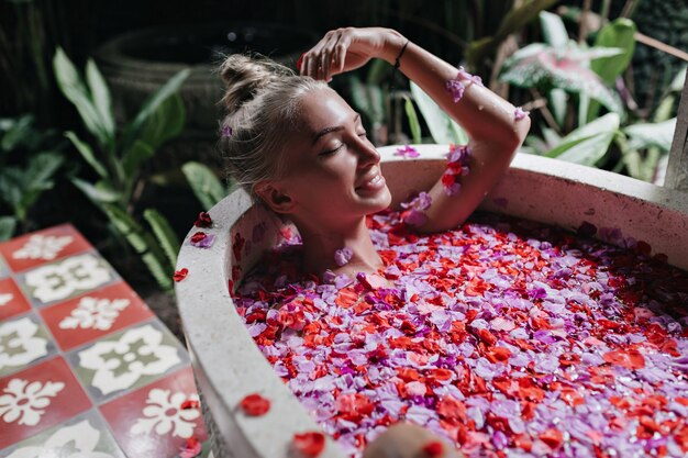 chica rubia sonriendo mientras se relaja en el baño. Hermosa mujer caucásica divirtiéndose durante el spa con flores.