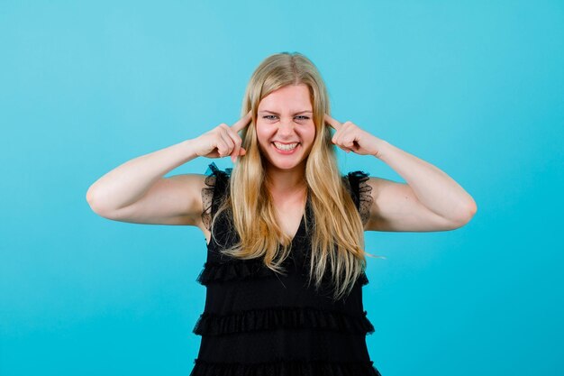 Chica rubia sonriente está cubriendo sus oídos con los dedos índices sobre fondo azul.