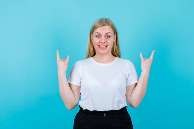 Chica rubia sonriente está haciendo gestos de rock sobre fondo azul.