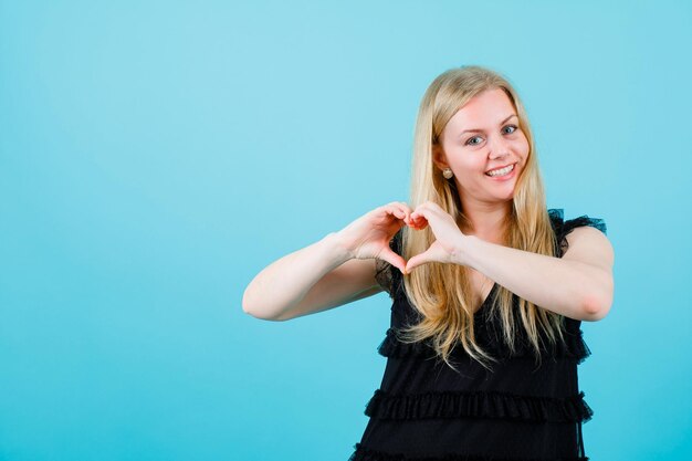 Chica rubia sonriente está mostrando un gesto de corazón sobre fondo azul.