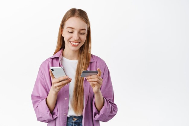 Chica rubia sonriente pagando compras en línea en la aplicación de teléfono inteligente y mirando su tarjeta de crédito de pie contra el fondo blanco