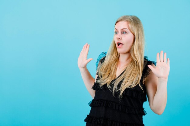 Chica rubia sorprendida está levantando sus puñados sobre fondo azul.