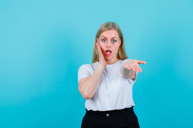 Chica rubia sorprendida está poniendo una mano en la mejilla y extendiendo la otra a la cámara sobre fondo azul.