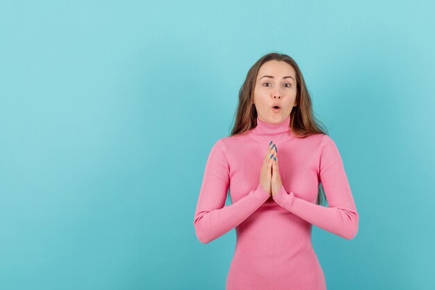 Chica rubia sorprendida está rezando tomándose de la mano en el fondo azul