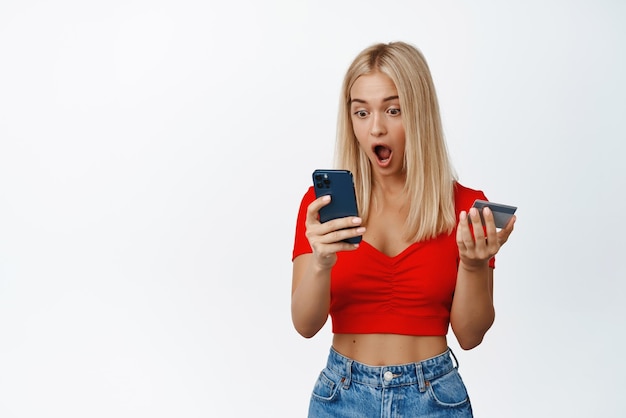 Chica rubia sorprendida mirando la pantalla del teléfono inteligente mientras hace un pedido navegando por el sitio de compras en el teléfono móvil con fondo blanco de la tarjeta de crédito