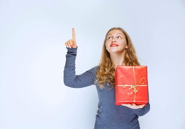 Chica rubia sosteniendo una caja de regalo roja y apuntando hacia arriba.