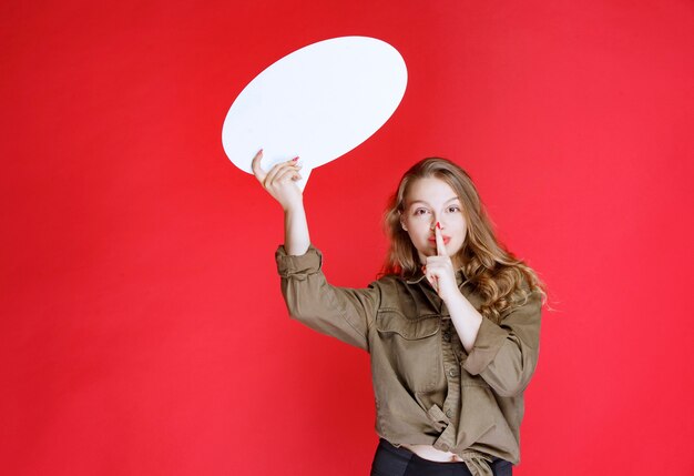 Chica rubia sosteniendo un thinkboard ovale y pidiendo silencio.