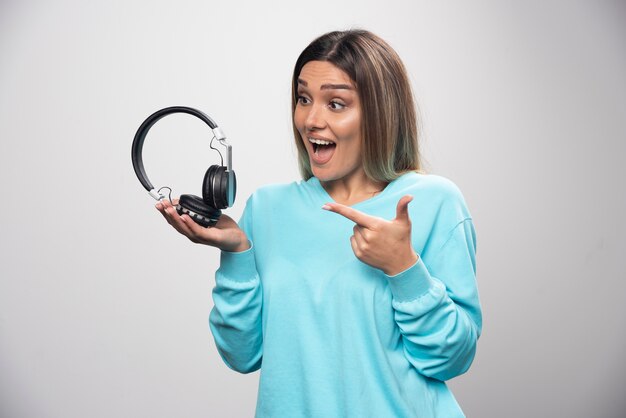 Chica rubia en sudadera azul sosteniendo auriculares y se prepara para usarlos para escuchar música