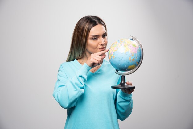 Chica rubia en sudadera azul sosteniendo un globo terráqueo, adivinando la ubicación y divirtiéndose.