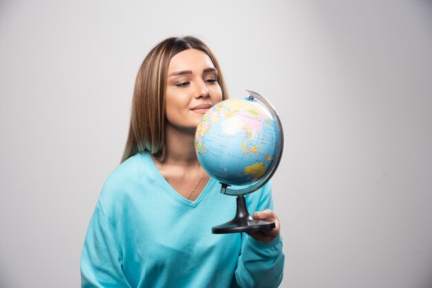 Chica rubia en sudadera azul sosteniendo un globo terráqueo, adivinando la ubicación y divirtiéndose.