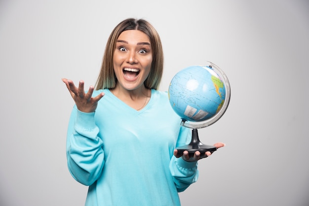 Chica rubia en sudadera azul sosteniendo un globo terráqueo, adivinando la ubicación y divirtiéndose.