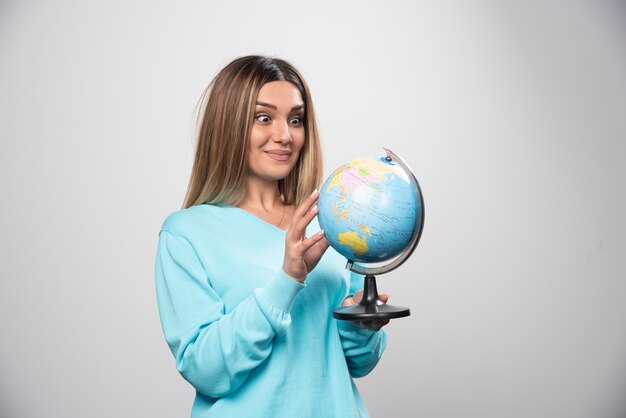 Chica rubia en sudadera azul sosteniendo un globo terráqueo, adivinando la ubicación y divirtiéndose.