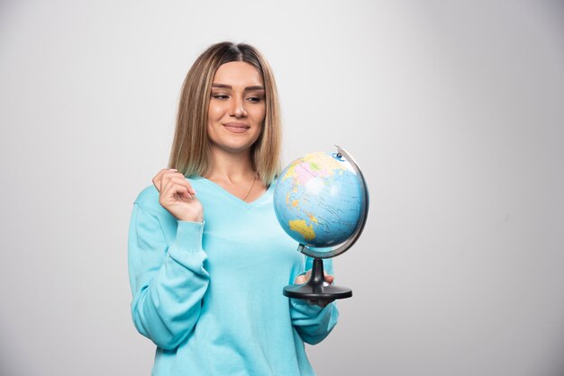 Chica rubia en sudadera azul sosteniendo un globo terráqueo, adivinando la ubicación y divirtiéndose.