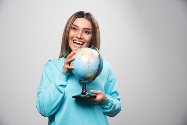 Chica rubia en sudadera azul sosteniendo un globo terráqueo, adivinando la ubicación y divirtiéndose