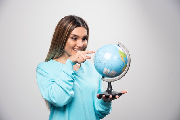 Chica rubia en sudadera azul sosteniendo un globo terráqueo, adivinando la ubicación y divirtiéndose