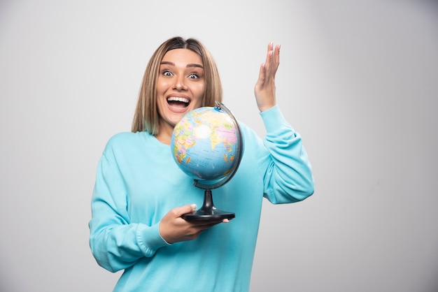 Chica rubia en sudadera azul sosteniendo un globo terráqueo, adivinando la ubicación y divirtiéndose