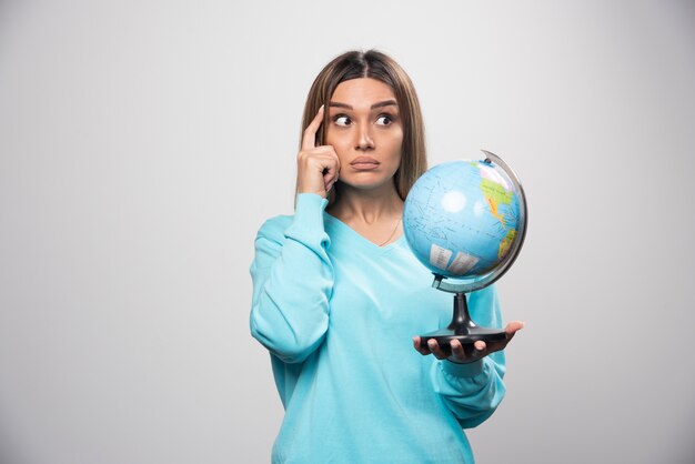 Chica rubia en sudadera azul sosteniendo un globo terráqueo, pensando cuidadosamente y tratando de recordar.