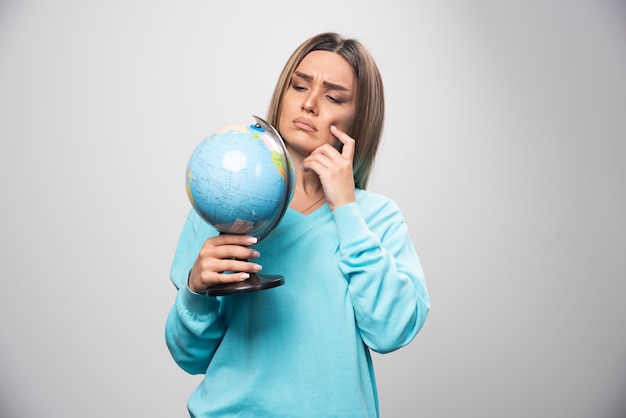 Chica rubia en sudadera azul sosteniendo un globo terráqueo, pensando cuidadosamente y tratando de recordar.