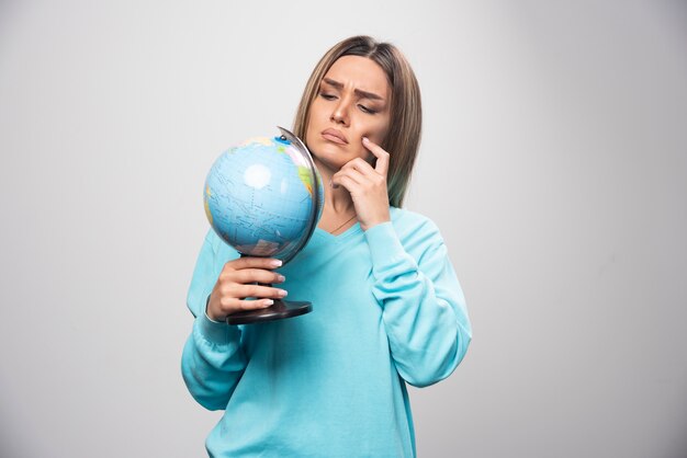 Chica rubia en sudadera azul sosteniendo un globo terráqueo, pensando cuidadosamente y tratando de recordar