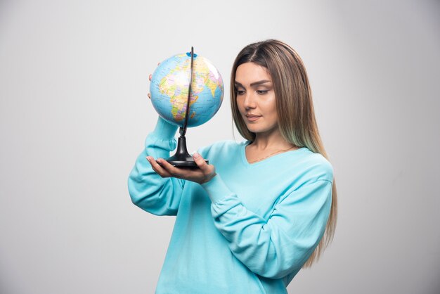 Chica rubia en sudadera azul sosteniendo un globo terráqueo y revisando el mapa de la tierra con atención.