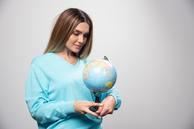 Chica rubia en sudadera azul sosteniendo un globo terráqueo y revisando el mapa de la tierra con atención.
