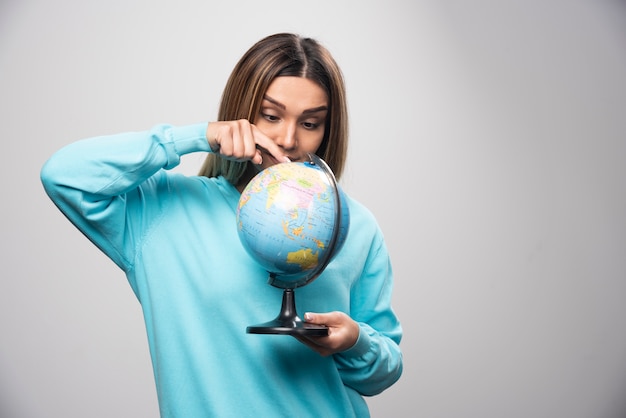 Chica rubia en sudadera azul sosteniendo un globo terráqueo y revisando el mapa de la tierra con atención