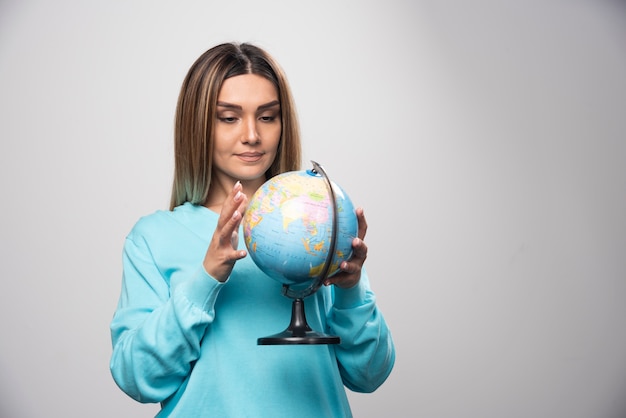 Chica rubia en sudadera azul sosteniendo un globo terráqueo y revisando el mapa de la tierra con atención