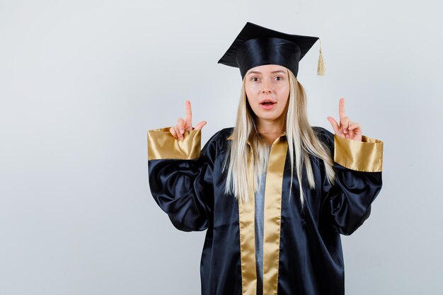 Chica rubia en toga y gorra de graduación apuntando hacia arriba con los dedos índices y luciendo linda